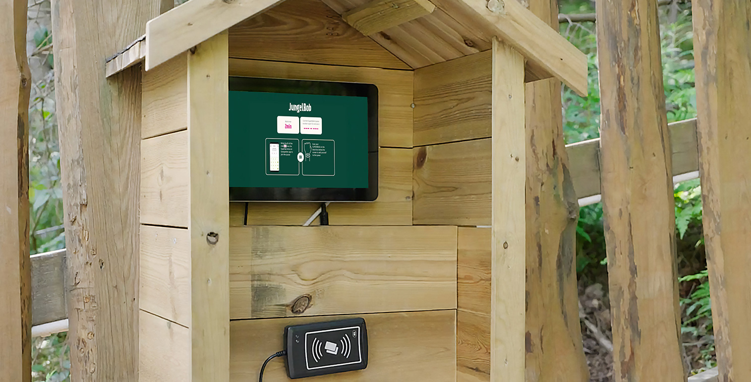 A visitor interacts with a weatherproof touchscreen kiosk housed in a wooden enclosure at an amusement park, showcasing self-service technology for ticketing.
